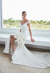 Scottie wedding dress by Morilee featuring an off-the-shoulder neckline, twist draped bodice, and lace-trimmed slit on a sleek sheath silhouette