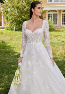 Morilee Sannah wedding dress style 2750 featuring a strapless sweetheart neckline, Alençon lace bodice, satin bow waistband, long sleeves, and scalloped hem tulle skirt